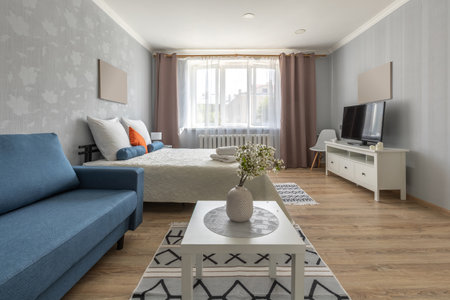 A bright studio-style room featuring a cozy bed with rolled towels, a blue sofa, white coffee table with vase, TV stand, and wardrobe under a large sheer-curtained window.の写真素材