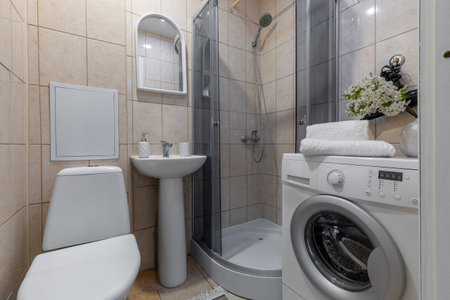 A compact bathroom featuring a pedestal sink, corner shower, and front-loading washing machine, with decorative flowers, towels, and beige tiled walls creating a clean and functional space.の写真素材