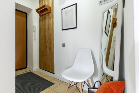 Modern entryway featuring a wooden wall panel, white chair, leaning mirror, framed artwork, and a telephone near the door.の写真素材