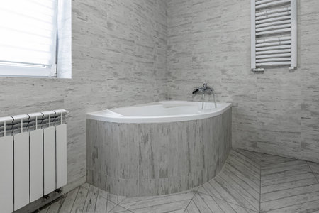 Clean corner bathtub with light gray tile finish, radiator and towel dryer in a modern minimalist bathroom.の写真素材
