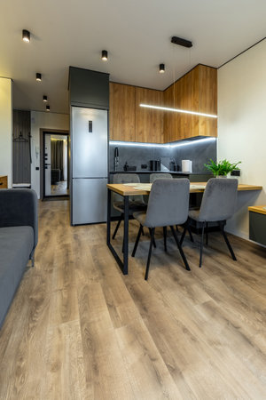 Stylish kitchen with wood cabinets, gray backsplash, dining table with soft chairs, and warm lighting in a cozy interior.の写真素材