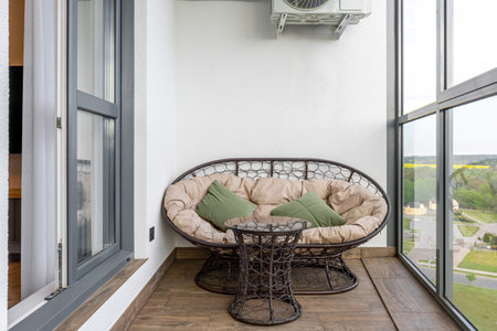 Small glazed balcony with round rattan sofa, glass-top table, soft cushions, and panoramic view of nature and buildings.の写真素材