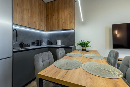 Compact modern kitchen with wooden cabinets, gray backsplash, soft chairs, and a large wooden dining table with placemats and a plant.の写真素材