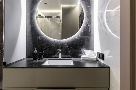 A sleek modern bathroom vanity featuring a black marble backsplash, integrated sink, backlit round mirror, and stylish accessories for a luxurious spa-like feel.の写真素材