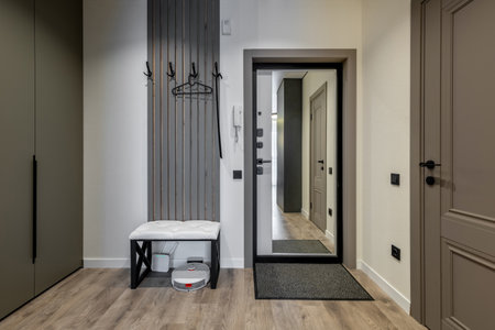 Modern entryway featuring wall hooks, upholstered bench with storage, full-length mirror, and robotic vacuum on laminate wood-patterned floor.の写真素材