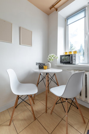 A cozy kitchen corner with a round white table, modern chairs, a floral vase and microwave under natural window light.の写真素材