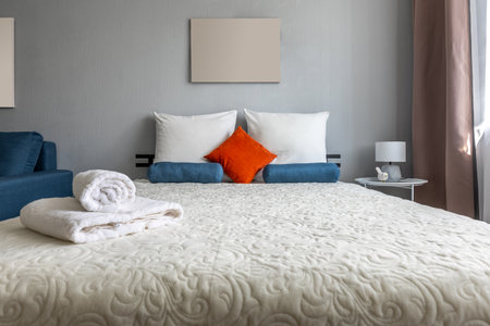 Rolled white towels are placed atop a plush quilted bed with blue bolsters and an orange accent pillow beside a sheer curtained window and bedside lamp..の写真素材