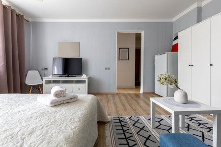 A bright multipurpose room combines a quilted bed with white towels, a TV stand, blue ottoman,...