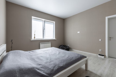 Bright minimalist bedroom with neutral tones, large window with blinds, white radiator, and a low white bed with gray bedding.の写真素材