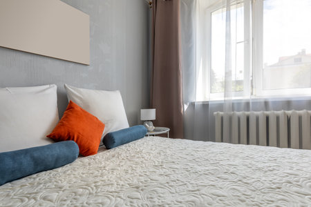 A brightly lit bedroom featuring quilted bedding with blue bolsters and an orange accent pillow beside a sheer-curtained window and bedside lamp.の写真素材