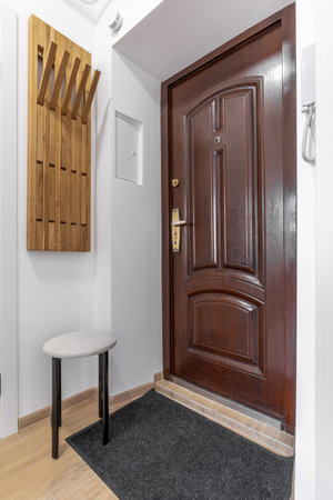 Interior view of a closed brown wooden entrance door in a modern white hallway with a wall-mounted coat rack and small round stool.の写真素材