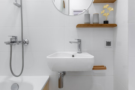 Close-up of a modern white sink and chrome shower in a minimalist bathroom with wooden shelf and elegant decor.の写真素材