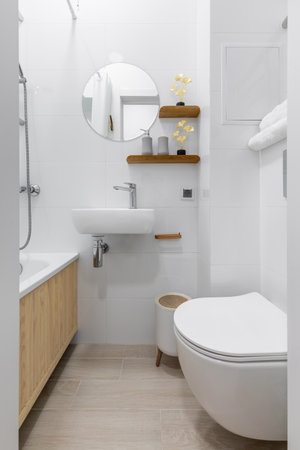 Bright modern bathroom with white tiles, wall-mounted toilet, compact sink, round mirror, and wooden accent shelves with decor.の写真素材