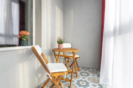 Bright cozy balcony with wooden chairs, small round table, patterned tile floor, and soft natural...
