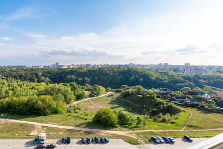 Aerial cityscape with green forest, houses, parked cars, and distant residential buildings under a clear blue sky.の写真素材