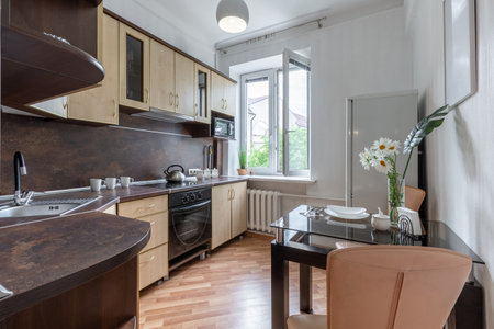 Modern bright kitchen interior with wooden cabinets, glass dining table, open window and cozy...