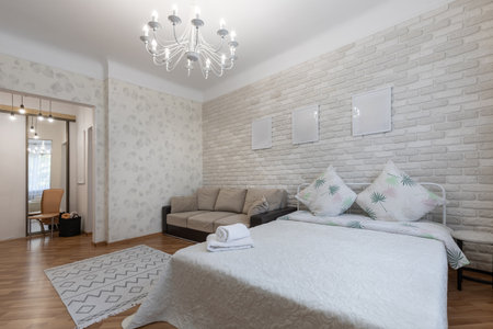 Spacious bright bedroom interior with white brick wall, cozy bed, beige sofa, elegant chandelier and...