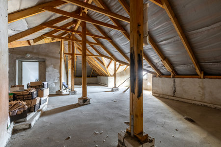 Spacious unfinished attic with wooden roof supports, rough concrete floor, plastered walls, and scattered storage boxes in a construction-stage interior.の写真素材