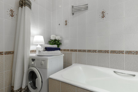 Bright bathroom interior featuring a bathtub, washing machine, lamp, towels, and decorative tiles.の写真素材