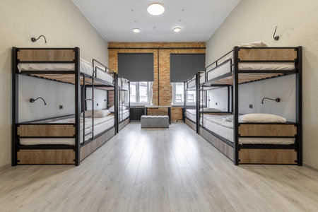 A perfectly symmetrical shot of a clean hostel dormitory featuring four black metal bunk beds with wood panels. The room includes authentic yellow brick walls between two windows, grey roller blinds, and bright laminate flooring.の写真素材
