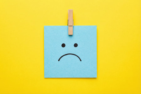 Sad face on blue note paper with clothespins on yellow background.の素材