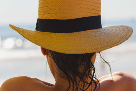 Beautiful young girl wearing a yellow straw hat at the beach, view from behind. Hat with black cloth...