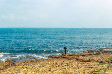 Fisherman fishing in Oeiras, Portugal. Deep blue ocean and rocky beach landscape, with shallow...