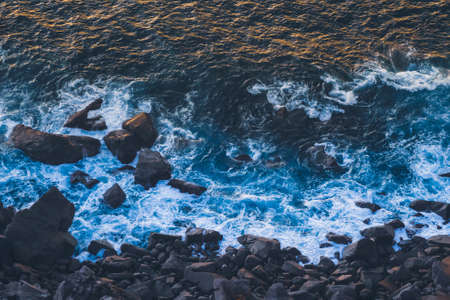 Deep blue breaking waves of atlantic ocean hitting on dark basalt shore with golden sunlight reflecting off waves at sunset. Cape Rocaの写真素材