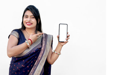 A Portrait of happy beautiful indian female wearing saree holding a smart phone with white or blank screen, promoting app, advertisement, isolated on white backgroundの写真素材