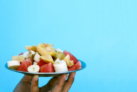 Close-up of a hand holding a metal plate with freshly cut fruit pieces including banana and...