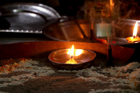 A traditional oil lamp (diya) burning with a bright flame placed on sacred sand during a Hindu ritual, with incense smoke and offerings in the backgroundの写真素材