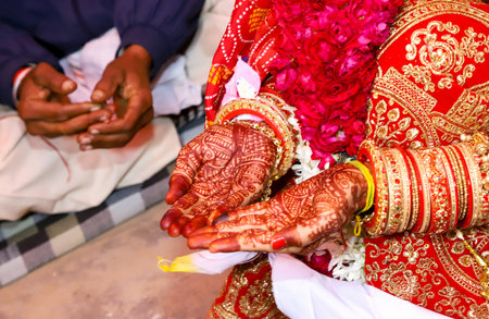Close-up of an Indian brideâs hands adorned with intricate mehndi designs, red and golden bangles, floral garland, and traditional embroidered red bridal attire during a wedding ceremony.の写真素材