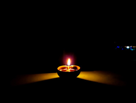 A traditional clay oil lamp (diya) with a burning cotton wick glowing softly in the dark,...
