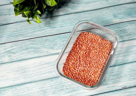 A clear glass container filled with red lentils (masoor dal) placed on a light wooden surface, showcasing clean, dry pulses for cooking or food photography.の写真素材