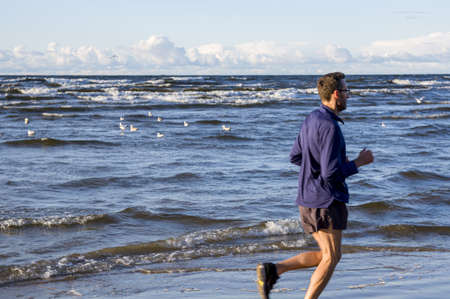 Exercising outside. A jogger in lightweight blue long sleeve t-shirt, shorts and running sneakers trains to marathon, running across the Baltic Sea beach, on cloudy windy early morningの写真素材