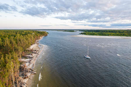 View from above, stunning aerial view of sailing white luxury modern not big sail boat or yacht with folded sail at an estuary of blue river enters sea next to green coastline at golden hour at sunsetの写真素材