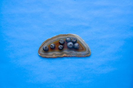 Flat lay of red agate gemstone and juicy, fresh blueberries on blue table. Isolated sweet bilberry...
