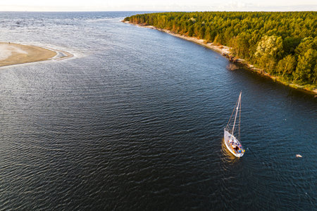 View from above, stunning aerial view of sailing white luxury modern not big sail boat or yacht with folded sail at an estuary of blue river enters sea next to green coastline at golden hour at sunsetの写真素材