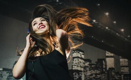 A studio shot of a beautiful brunette woman wears purple headphones playing with her hair, listening music and dnacing before city wallpaperの写真素材