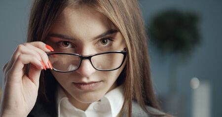 Close-up of beautiful confident business girl wearing glasses holding frame and looking at camera over the top of specsの写真素材
