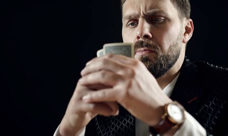 Close-up of mature bearded man in suit gambling holding cards in front of his face in dark...