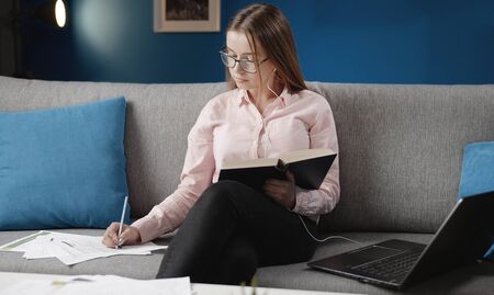 Nice female student wearing glasses and earphones having open book in her hand and making notes...