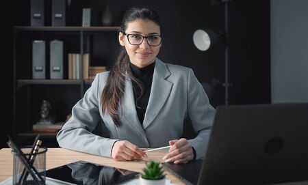 Glad businesswoman in eyeglasses and formal dress looking at camera while sitting at workplace in officeの写真素材