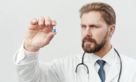 Serious bearded doc with stethoscope holding one blue and white capsule between his fingers looking...