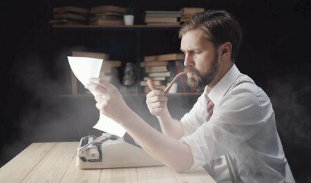 Serious bearded novelist looking at paper checking typescript and smoking pipe, bookcase backgroundの写真素材