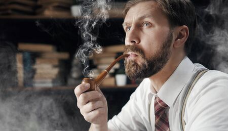 Portrait of thoughtful bearded mature man smoking wooden pipe over bookshelf backgroundの写真素材