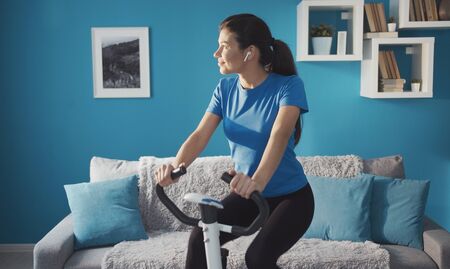 Sporty happy girl training on exercise bike at home listening music looking aside, healthy lifeの写真素材