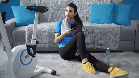 Girl listen to music on smartphone and rest sitting on floor after training on cycling machine, homeの写真素材