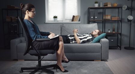 Man lying on sofa speaking about his problems with woman psychologist sitting in chair making notesの写真素材