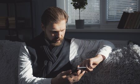 Middle-aged bristled man of respectable appearance using smartphone sitting on sofa at homeの写真素材